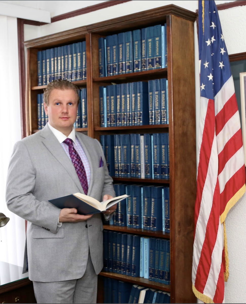 New York attorney Michael L. Walker reading a legal volume beside the NY Statutes library and American flag at his Bay Ridge, Brooklyn office
